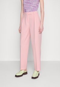 Un pantalon ajusté rose clair à taille haute, avec des plis et une coupe décontractée, associé à des mocassins vert menthe sur une semelle marron foncé.