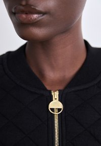 Jaqueta bomber acolchoada preta com um puxador de zíper dourado, apresentando um colarinho arredondado e um tecido texturizado.