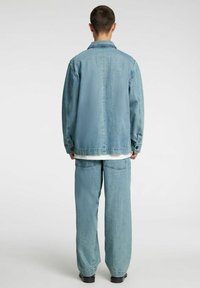 Lichtblauwe spijkerjasje met een kraag, gecombineerd met losvallende lichtblauwe spijkerbroeken. Beide hebben een gestructureerde afwerking en zichtbare stiksel details.