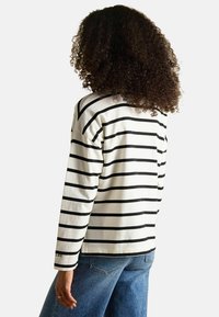 Langärmliges gestreiftes Oberteil in Creme und Schwarz, mit lockerer Passform und gerundetem Saum. Sichtbare Logodetails in der Nähe des Saums. Getragene Denim-Jeans.