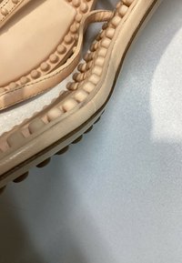 Gros plan sur des sandales beige avec des clous ronds texturés le long des brides et des bords de la semelle sur une surface claire.