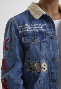 Denim jacket with a shearling collar, blue color, featuring embroidered text, red felt patches, and large numerical accents in gold and beige.