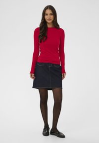 Frau steht mit einem roten Langarmtop, einem dunklen Jeansrock, schwarzen Strumpfhosen und schwarzen Loafers vor einem schlichten hellen Hintergrund.