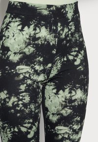 Svarta leggings med ett grönt tie-dye-mönster, texturerad tygkvalitet och en slät midjeresår. Närbild framhäver designens detaljer.