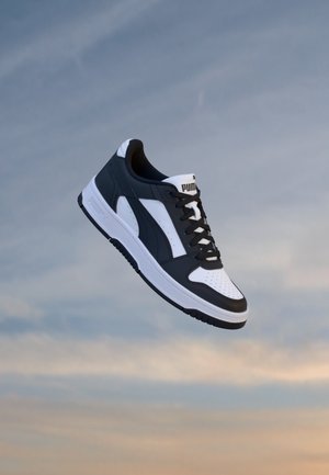 Baskets Puma noir et blanc flottant sur un fond de ciel bleu et orange.