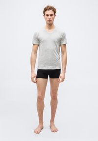 Jeune homme debout, pieds nus, portant un T-shirt gris clair à col en V et un boxer noir, sur un fond clair uni.