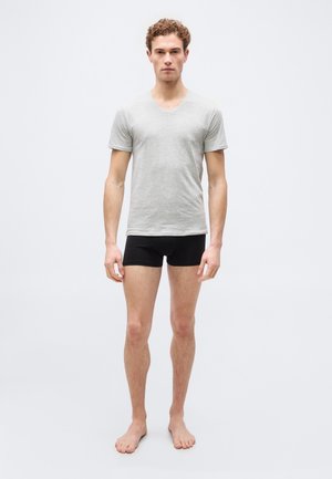 Jonge man staat op blote voeten, draagt een lichtgrijs T-shirt met V-hals en zwarte boxershorts tegen een effen lichte achtergrond.