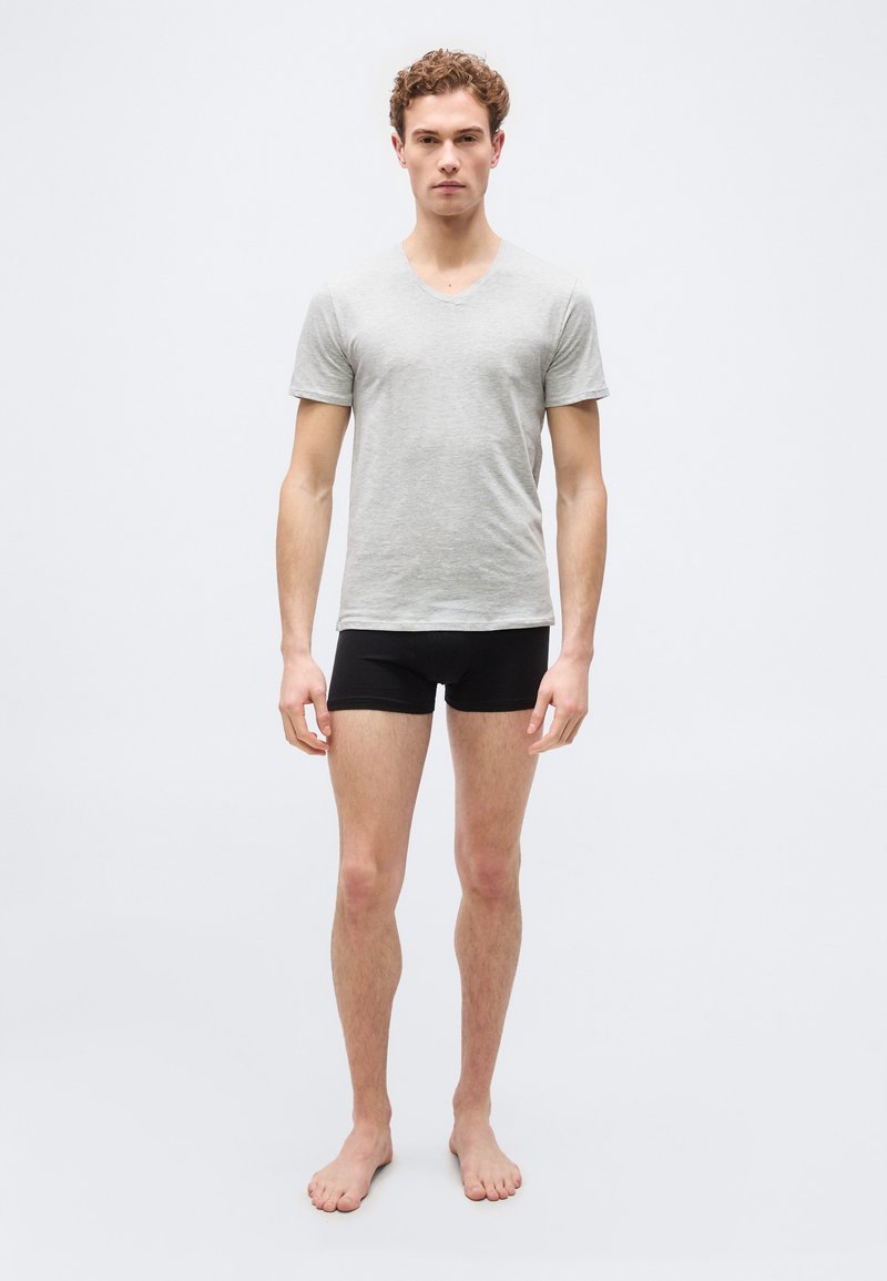 Jeune homme debout, pieds nus, portant un T-shirt gris clair à col en V et un boxer noir, sur un fond clair uni.