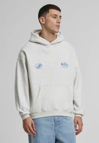 Hanorac cu glugă de culoare gri deschis, cu buzunar frontal; prezintă un logo imprimat albastru "A.C.L." și un accent grafic pe partea stângă. Textură moale.