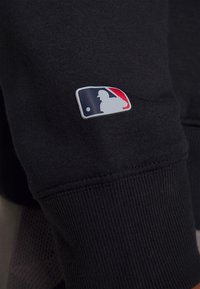 Sweat-shirt noir avec un patch logo MLB blanc et coloré sur la manche. Le tissu est lisse avec des poignets côtelés.