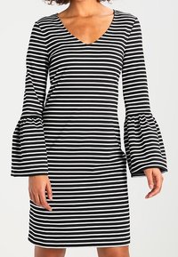 Mustavalkoinen raitapuku V-kaula-aukolla, jossa on pitkät kellohihat, tiukka vartalo ja pehmeä kangas.