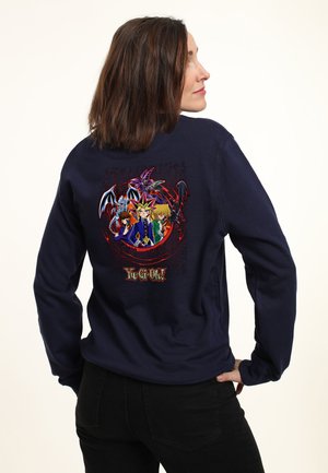 Kvinde i marineblå sweatshirt med farverige Yu-Gi-Oh-figurer og logo trykt på ryggen, står med hånden på hoften og kigger sidelæns.