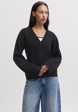 Donna con lunghi capelli scuri che indossa un cardigan nero con bottoni sopra una maglietta a righe e jeans azzurri, in piedi davanti a uno sfondo semplice.