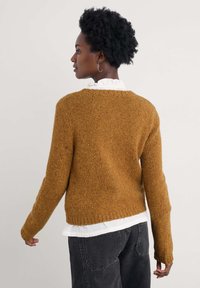 Mosterdgele gebreide trui met een ronde hals en geribbelde zoom. Gedragen over een witte top met ruffles, die lange mouwen heeft en een textuurfinish.