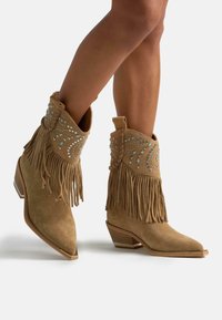 Botas de cowboy de ante marrón con flecos y decorativas piedras brillantes; diseño de punta afilada y tacón apilado, mostrando una superficie texturizada.