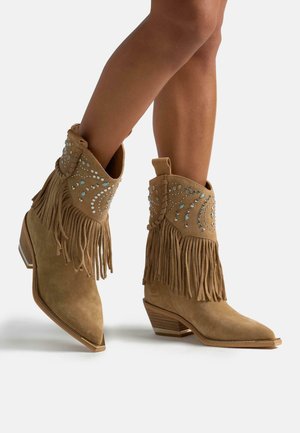 Botas de cowboy de ante marrón con flecos y decorativas piedras brillantes; diseño de punta afilada y tacón apilado, mostrando una superficie texturizada.