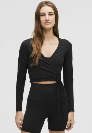 Ung kvinde med langt brunt hår iført en sort omlagt langærmet cropped top og højtaljede sorte shorts, stående mod en ensfarvet baggrund.