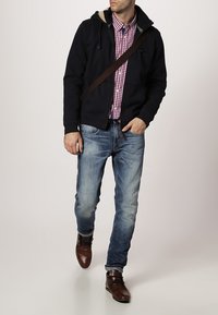 Uomo che cammina, indossando un felpa nera con cappuccio sopra una camicia a scacchi rossi, jeans blu, scarpe di pelle marroni e una borsa a tracolla marrone.