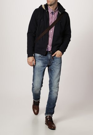 Uomo che cammina, indossando un felpa nera con cappuccio sopra una camicia a scacchi rossi, jeans blu, scarpe di pelle marroni e una borsa a tracolla marrone.
