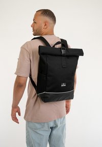 Rucsac negru cu capac rulabil, bretele ajustabile, buzunar frontal cu fermoar și detaliu argintiu la bază. Fabricat din material durabil.