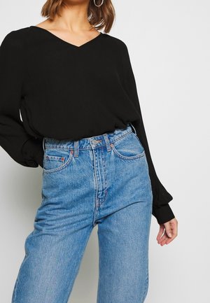 Femme portant un chemisier noir à manches longues en V, rentré dans un jean bleu clair taille haute, avec de grandes boucles d'oreilles anneaux argentées.