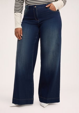 Person iført mørkeblå vidde jeans og en langærmet top med striber, med den ene hånd i lommen og hvide spidse sko.