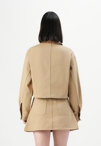 Kvinne med rett, svart hår, iført en beige kort jakke og matchende beige skjørt, står med ryggen mot en hvit bakgrunn.