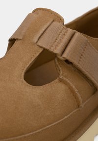 Bruine suède schoen met elastische band, ronde uitsnijding aan de zijkant, gladde textuur en platte zool met een contrasterende witte rand.