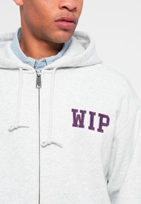 Gray zip-up hoodie with purple "WIP" embroidery on the chest. Features drawstrings and a layered blue collar beneath. Soft texture.