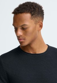 Zwart shirt met lange mouwen, voorzien van een geribbelde textuur en een ronde halslijn, met een close-up van de kraag en het bovenlichaam.