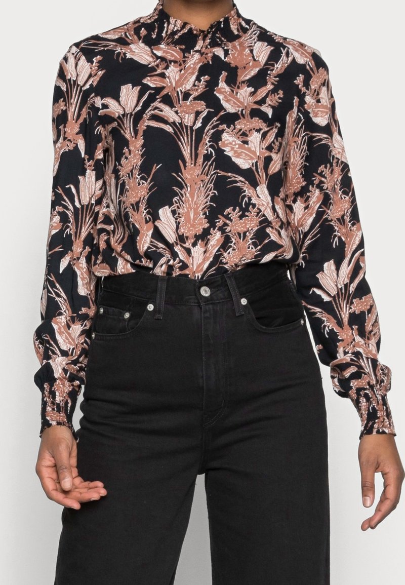 Jean noir taille haute combiné avec un chemisier noir à manches longues présentant un motif floral beige et des poignets froncés.