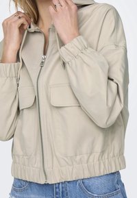 Beige zip-up jacket with a hood, elastic cuffs, and waistband; features two front pockets and a smooth, lightweight fabric texture.