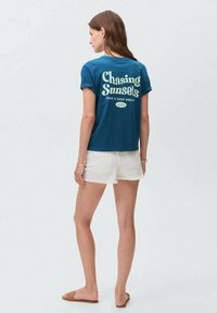 Tyrkysové bavlněné tričko s ohrnutými rukávy, na zadní straně je bílé nápis "Chasing Sunsets", spárované s bílými denimovými šortkami.