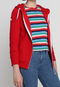 Rode hoodie met ritssluiting, witte trekkoorden en twee voorzakken, gedragen over een multicolor gestreept T-shirt en blauwe spijkerbroek.