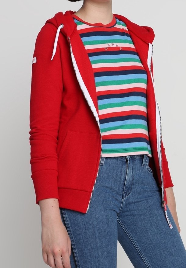 Rode hoodie met ritssluiting, witte trekkoorden en twee voorzakken, gedragen over een multicolor gestreept T-shirt en blauwe spijkerbroek.