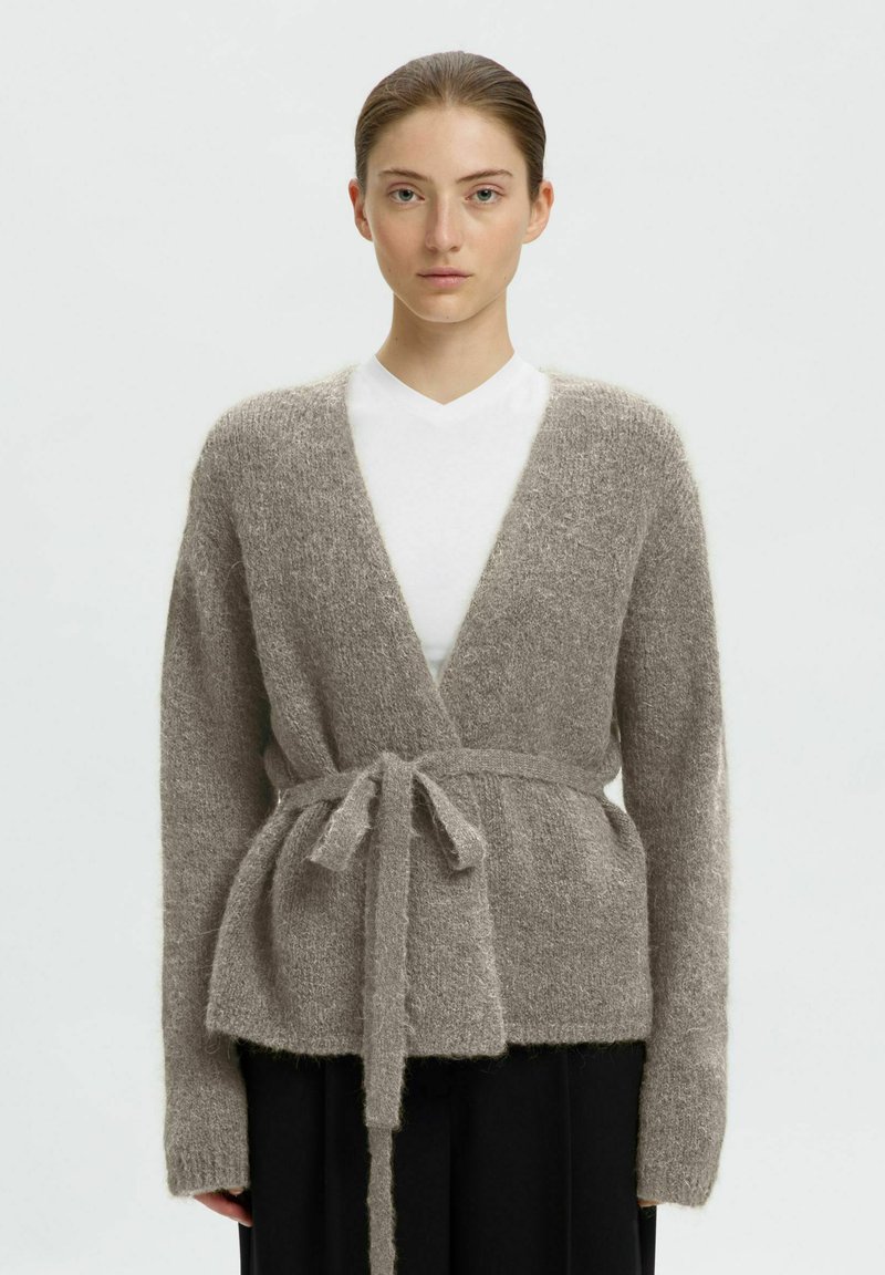 Jeune personne avec un cardigan gris duveteux noué sur une chemise blanche et un pantalon noir sur un fond clair uni.