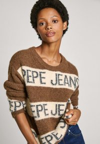 Maglione a righe marroni e crema realizzato in materiale morbido e fluffy. Presenta il testo "PEPE JEANS" in blu scuro, con un ampio scollo e maniche arrotolate.