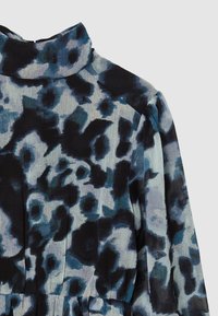 Hochkragen-Bluse mit langen Ärmeln, die ein blaues und schwarzes abstraktes Blumenmuster auf strukturiertem Stoff gegen einen einfarbigen Hintergrund zeigt.