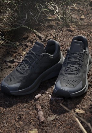 Par de zapatillas de running negras con cordones reposando sobre suelo seco con ramitas y hierba en un entorno natural al aire libre.