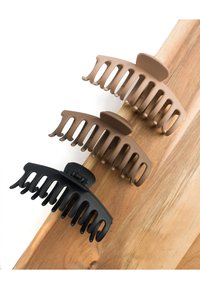 Drei Haarklammern aus schwarzem, hellbraunem und dunkelbraunem Plastik mit breiten Zähnen und einer abgerundeten Oberseite, angeordnet auf einer Holzoberfläche.