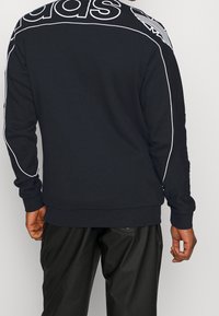 Sudadera negra con gran logo blanco de "adidas" en la parte posterior, con piping blanco en los hombros y puños acanalados.