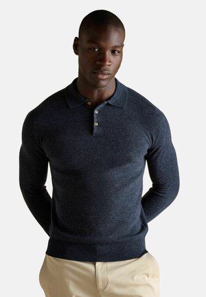 Marineblaues Strick-Polohemd mit einer Knopfleiste mit drei Knöpfen und gerippten Bündchen. Getragen mit beigen Hosen, die eine figurbetonte Silhouette aufweisen.