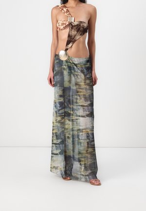 Kobieta ubrana w sukienkę maxi bez rękawów z abstrakcyjnym wzorem i metalicznymi okrągłymi ozdobami, nosząca sandały z odkrytymi palcami, stoi na białym tle.