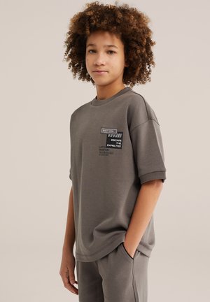 Junge Person mit lockigem Haar, die ein graues Kurzarmhemd und passende Hose trägt, das Hemd zeigt Text und einen Barcode-Grafik auf der Brust.