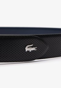 Cinturón de cuero perforado negro con logo de cocodrilo en metal plateado, diseño plano, textura suave y color interior contrastante.