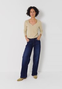 Mujer de pie con las manos en los bolsillos, con camiseta de manga larga beige, vaqueros anchos oscuros y zapatos beige puntiagudos sobre fondo blanco.