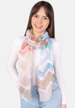 Junge Frau lächelt, trägt ein weißes Langarmshirt, hellblaue Jeans und einen bunt gemusterten Chevron-Schal um den Hals.