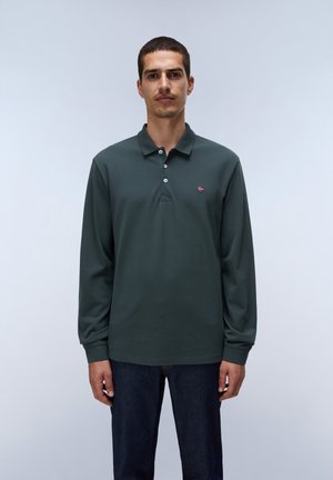 Mand med kort mørkt hår iført en mørkegrøn langærmet poloshirt og mørkeblå jeans, stående mod en ensfarvet lys baggrund.