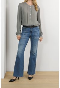 Blouse grise à manches longues avec fermeture à boutons, associée à un jean bleu évasé. Tenue complétée par des escarpins noirs à bout pointu et une ceinture noire.