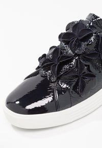 Baskets marines à patin avec accents floraux texturés, bout rond et semelle en caoutchouc blanche. Présente une finition brillante et des pétales doux et superposés.